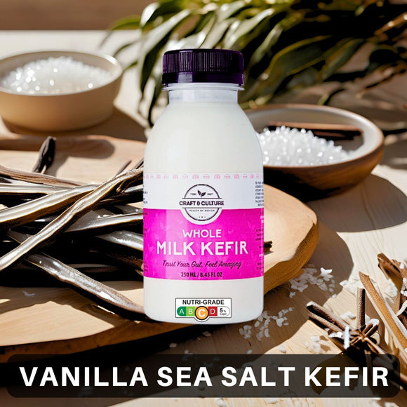 A bottle of [NEW] Vanilla Sea Salt Whole Milk Kefir from Craft & Culture - Kombucha, Kefir & Probiotics Singapore, adorned with a purple label, is placed on a table encircled by vanilla beans and bowls of coarse salt. At the bottom, the text 